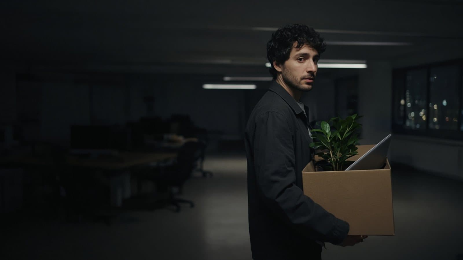 A man carries a cardboard box with a plant and a laptop through a dark office. Generated photograph. Not a picture of anyone in this story.